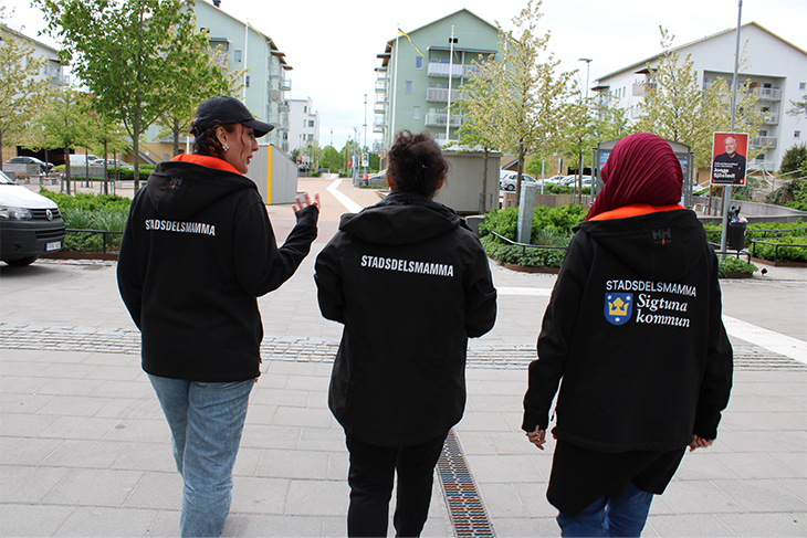 Tre kvinnor i svarta jackor med ordet stadsdelsmamma på ryggen går bredvid varandra i en stadsmiljö. 