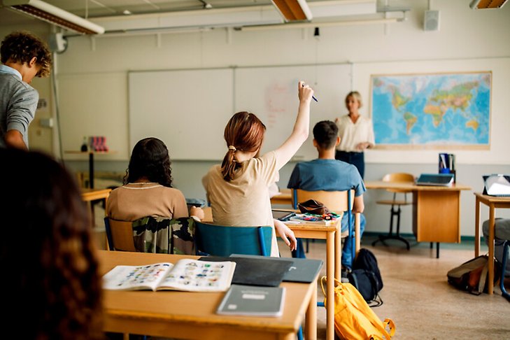 Elever sitter bakom sina bänkar i ett klassrum. En elev längst fram sträcker upp handen.