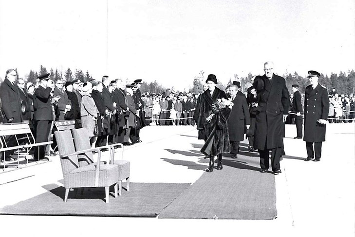 Kung Gustav VI Adolf och Drottning Louise på väg att sätta sig i två stolar framför publik.