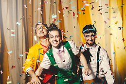 Tre glada personer i färgglada kläder står mitt i ett regn av konfetti