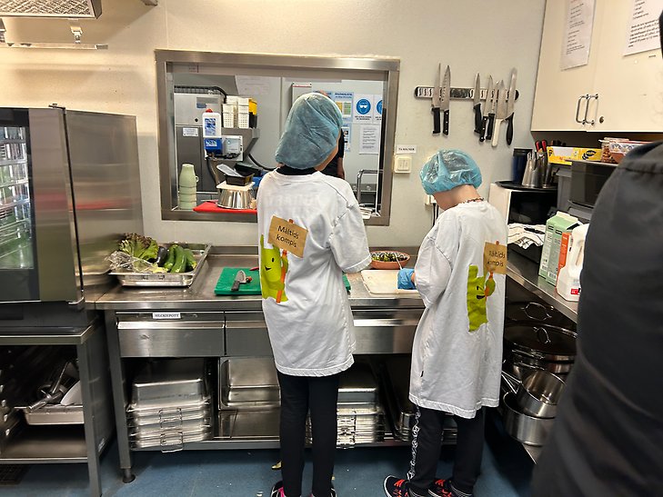 Två av deltagarna står vid en köksbänk i skolköket och hackar grönsaker. De står med ryggarna vända mot kameran, har blått hårnät på sig och en tröja där det står "Måltidskompis". Några av grönsakerna som ligger på arbetsbänken är zucchini, aubergine och sallad.